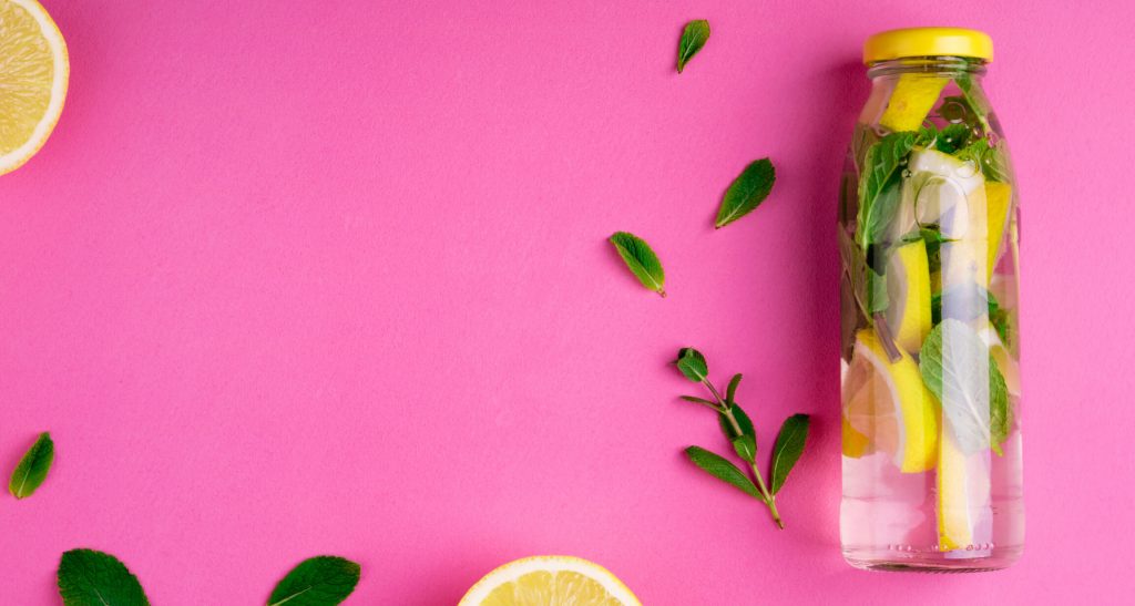 Glass water bottle filled with water, lemons, and herbs on a pink background